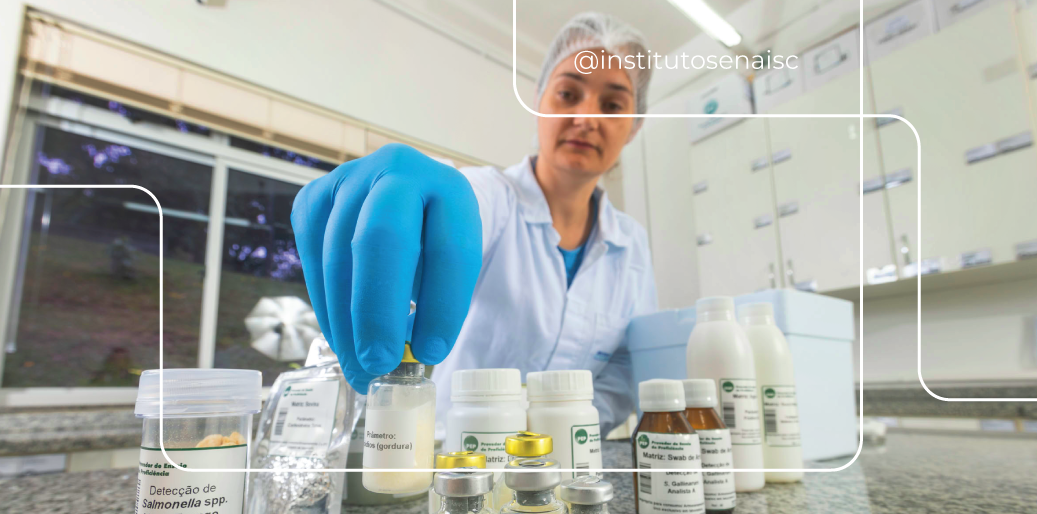Técnica de laboratório utilizando touca de proteção, jaleco branco e luvas azuis manipula frasco de material de referência em cima de uma bancada